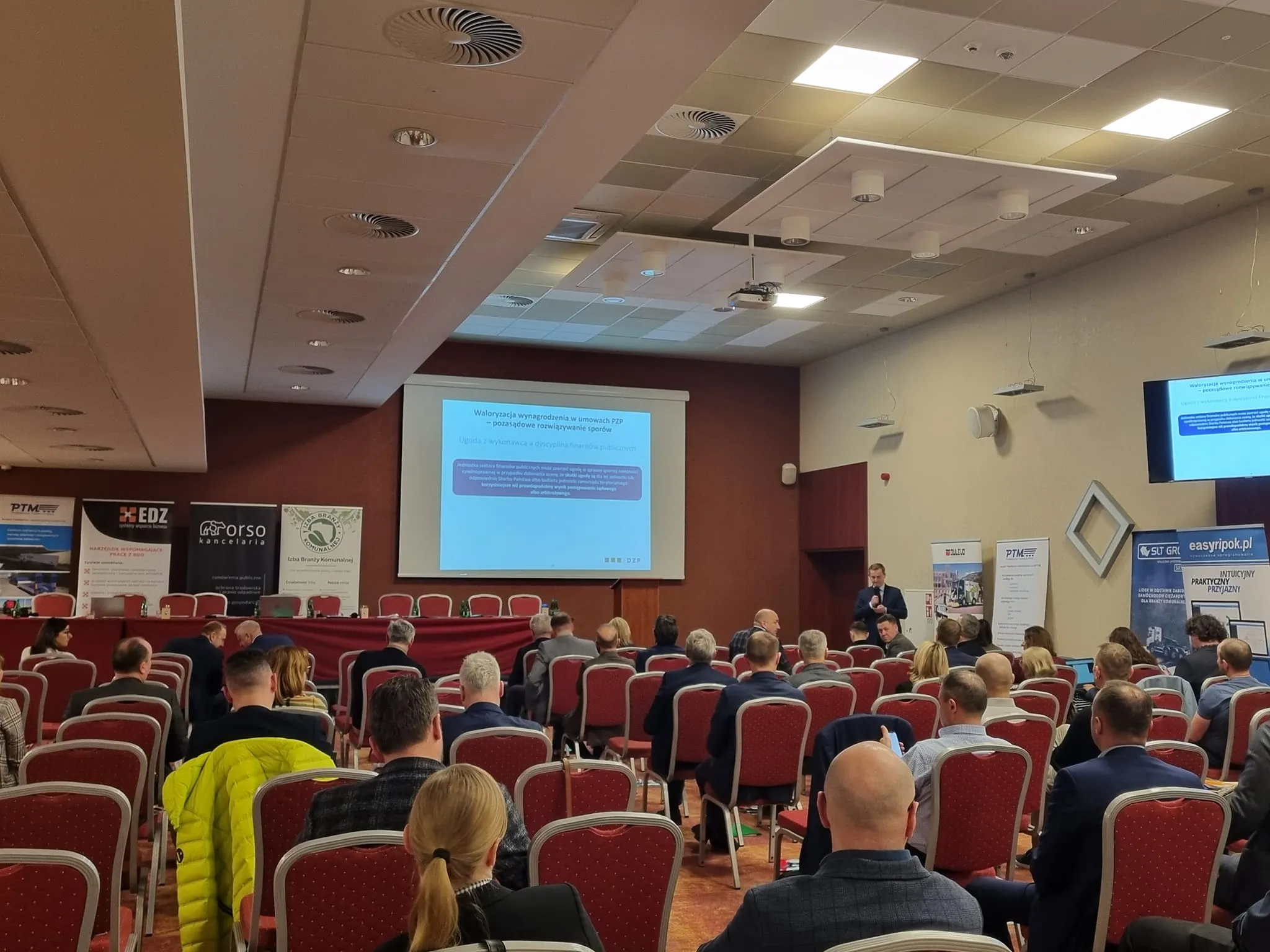 Uczestnicy I Zjazdu Szkoleniowo-Integracyjnego Izby Branży Komunalnej siedzą w sali konferencyjnej i słuchają prelekcji z prezentacją wyświetlaną na ekranie.