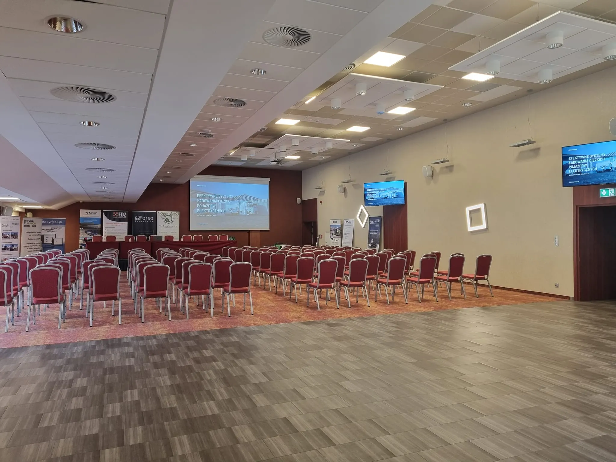 Sala konferencyjna przygotowana na I Zjazd Szkoleniowo‑Integracyjny Izby Branży Komunalnej z rzędami czerwonych krzeseł, stołem prezydialnym i ekranami prezentacyjnymi.