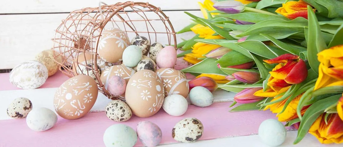 Wielkanocna kompozycja z kolorowymi pisankami wysypującymi się z koszyka i bukietem tulipanów na pastelowym tle.