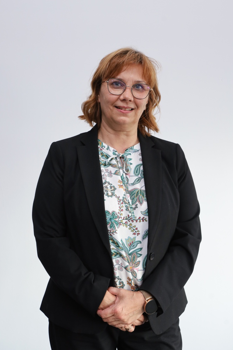 Kobieta w okularach w formalnym stroju (czarna marynarka, wzorzysta bluzka) pozuje na jasnym tle, reprezentując władze instytucji.