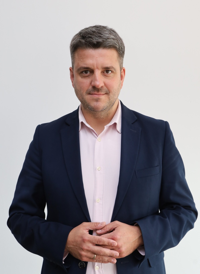 Mężczyzna w garniturze pozuje na neutralnym tle, przedstawiony w kadrze portretowym jako członek władz.