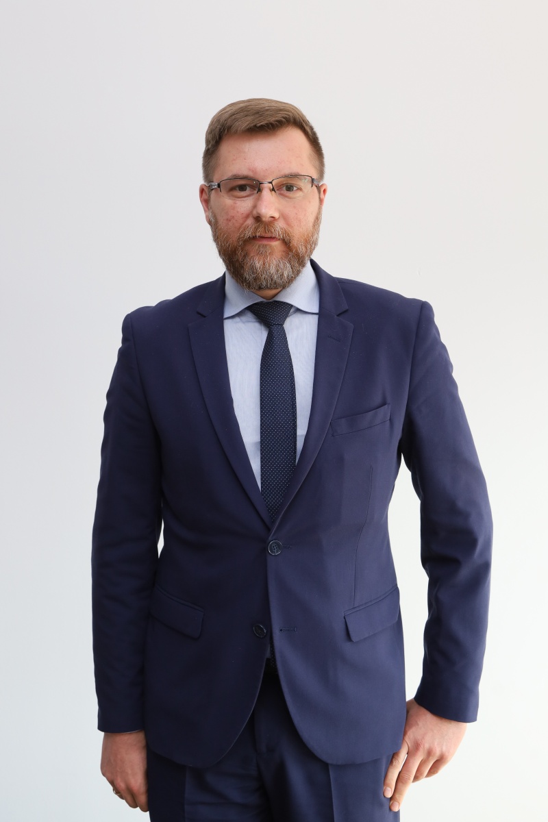 Mężczyzna w okularach ubrany w granatowy garnitur i krawat pozuje na jasnym tle, oficjalny portret przedstawiciela władz.