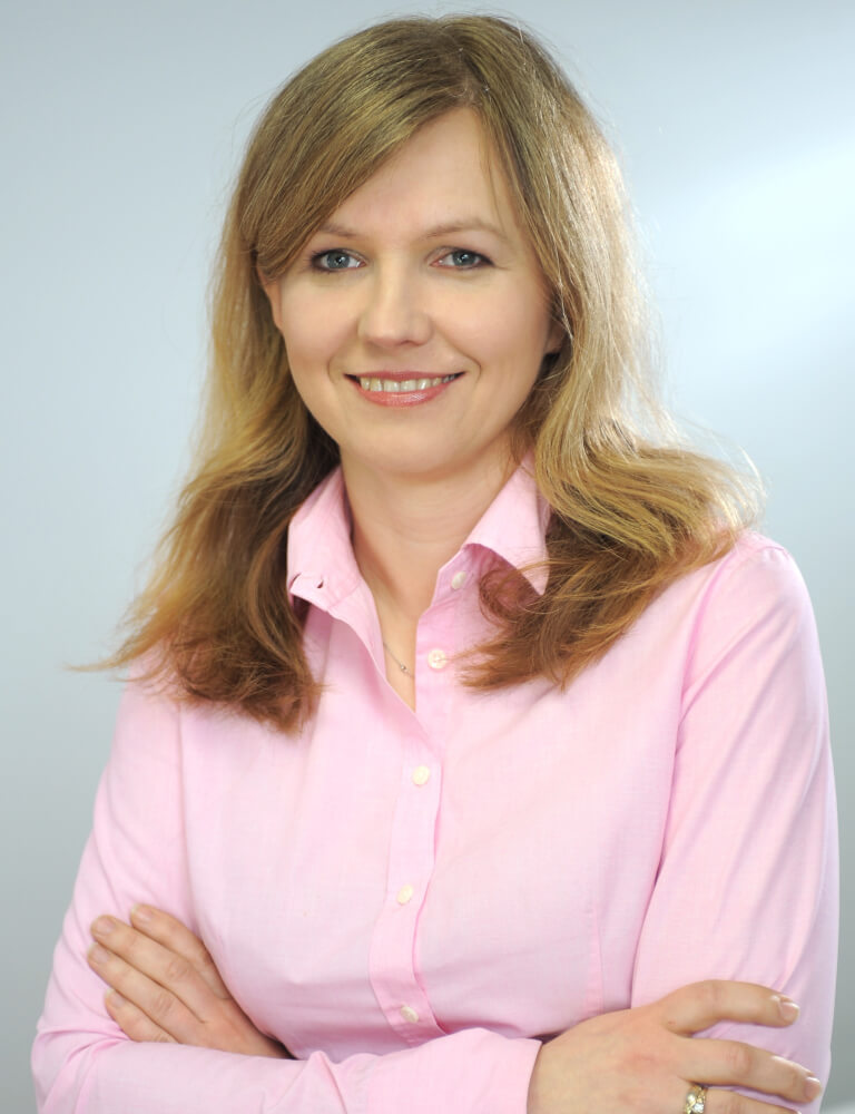 Portret uśmiechniętej kobiety w różowej koszuli, na neutralnym tle, z założonymi rękami.