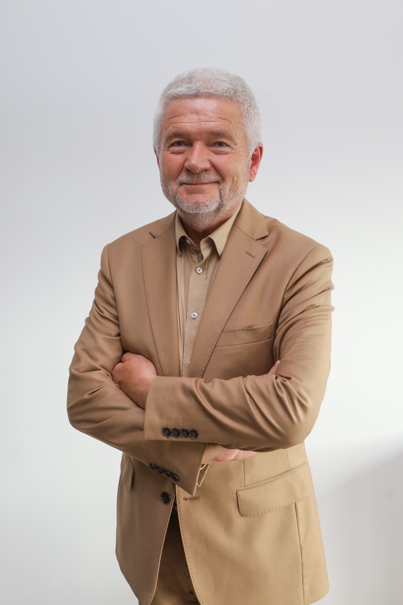 Mężczyzna w średnim wieku w beżowym garniturze pozuje z założonymi rękami na jasnym tle, portret władz instytucji.